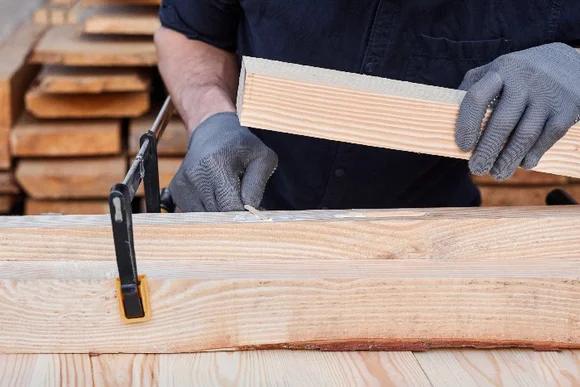 Person verklebt Holzbalken, die mit einer Schraubzwinge zusammengehalten werden